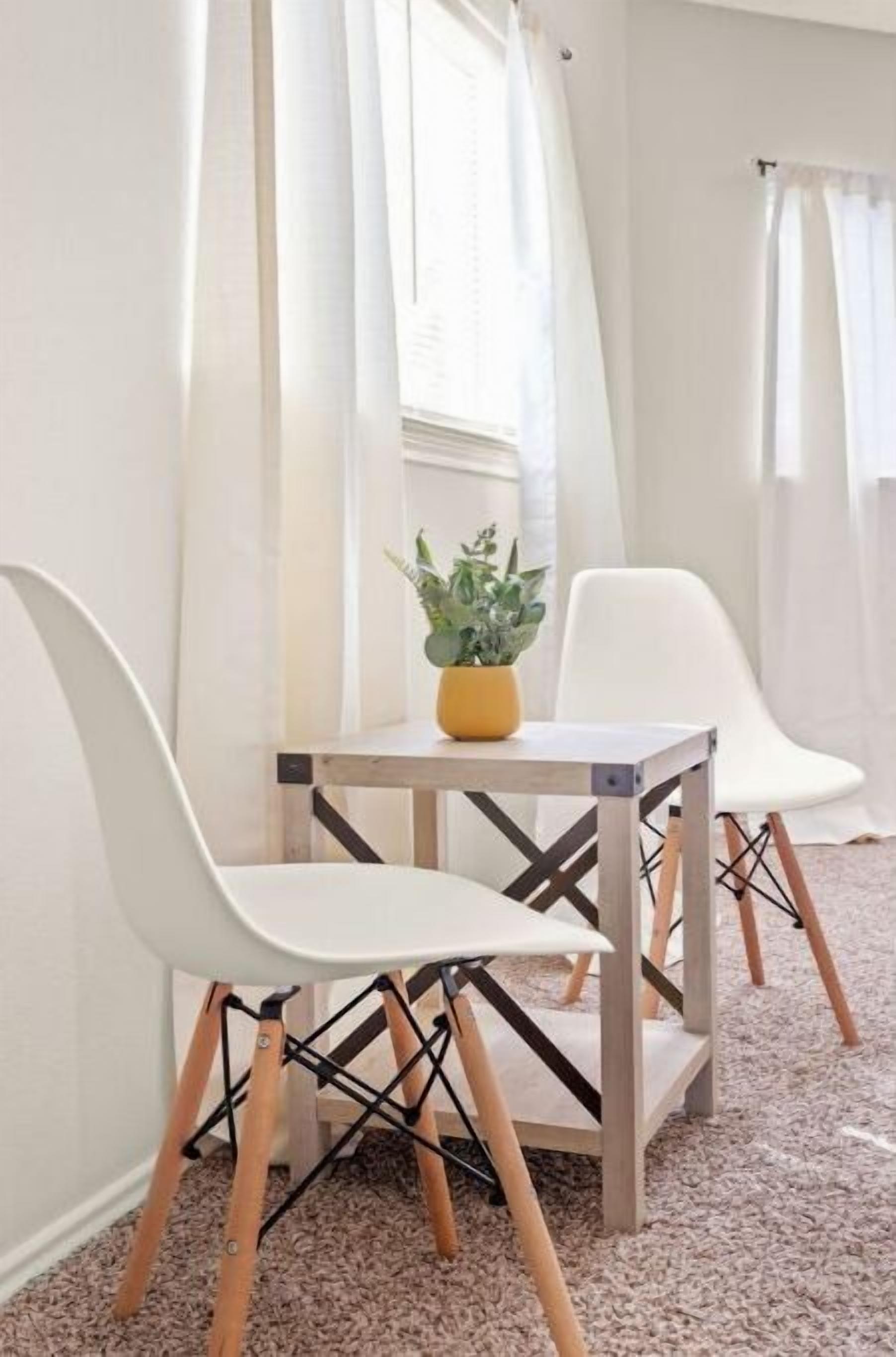 Ein Zimmer mit einem kleinen Tisch und zwei weißen Stühlen mit Holzbeinen. Auf dem Tisch steht eine Topfpflanze.