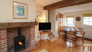 Ein gemütliches Wohnzimmer mit Kamin aus Ziegelsteinen, einem Fernseher auf einem Holzboden, einem Esstisch mit Stühlen und einem großen Fenster mit gelben Rollläden.