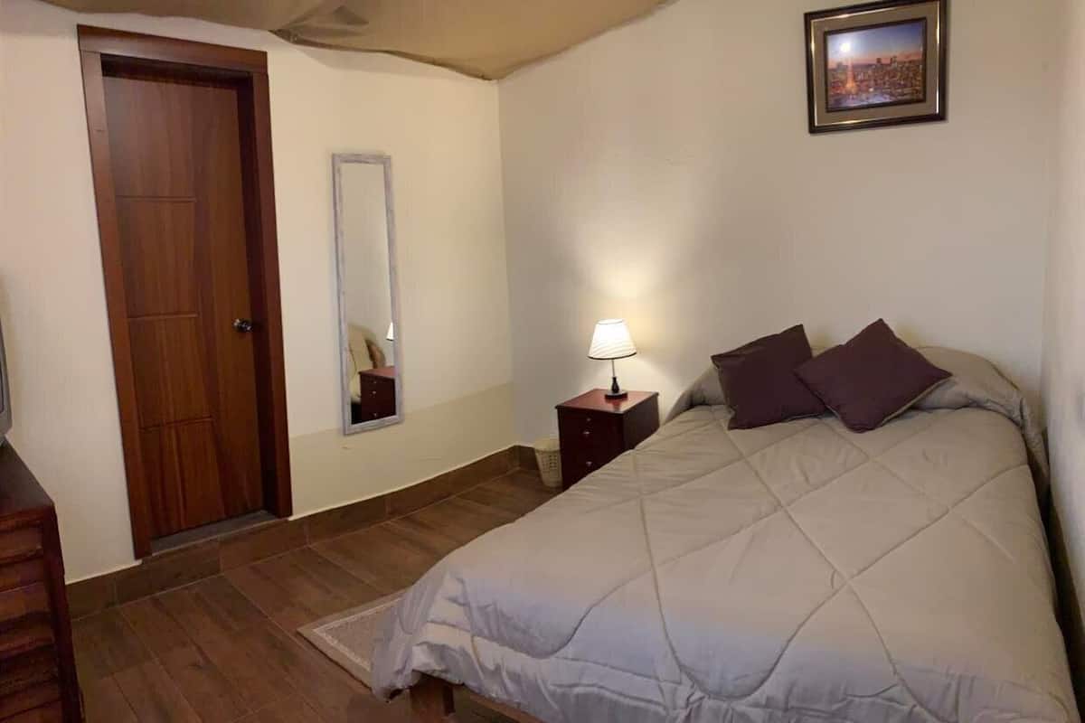 Un dormitorio con cama, mesita de noche, lámpara, puerta de madera y un cuadro en la pared.