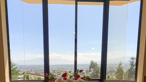 Une table à manger avec un plateau en verre et quatre chaises, un vase contenant des fleurs blanches et rouges, et une vue sur un paysage urbain par le biais d’une grande fenêtre.