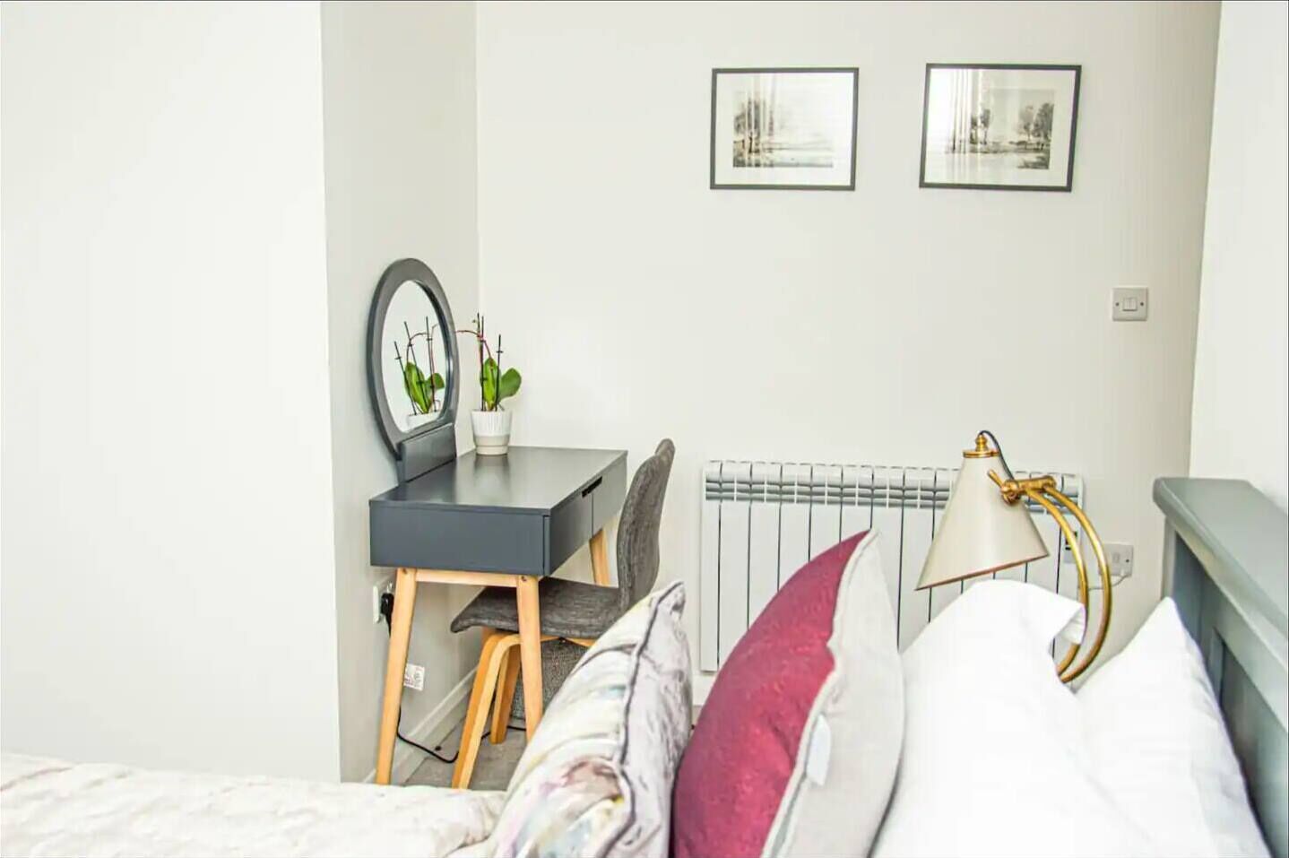 A bedroom with a bed, a desk, a chair, a radiator, and two framed pictures on the wall.
