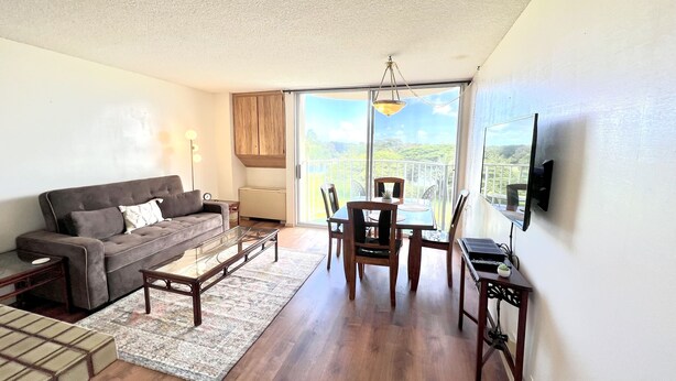 A living room with a sofa, coffee table, and dining table. There is a TV mounted on the wall and a window with a view of greenery. - Incredible View in Hilo - Pool, A/C, #708 (Hilo)