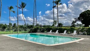 A rectangular swimming pool with a clear blue water surface, surrounded by a paved deck. There are four white lounge chairs aligned along the pool's edge. Tall palm trees are visible in the background, and the sky is partly cloudy. - Incredible View in Hilo - Pool, A/C, #708 (Hilo)