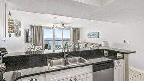 A modern kitchen with a black countertop, stainless steel sink, and dishwasher. There is a dining area with a table and chairs, and a view of the ocean through large windows. - Renovated Beachfront Condo with Sensational Views! (Destin)