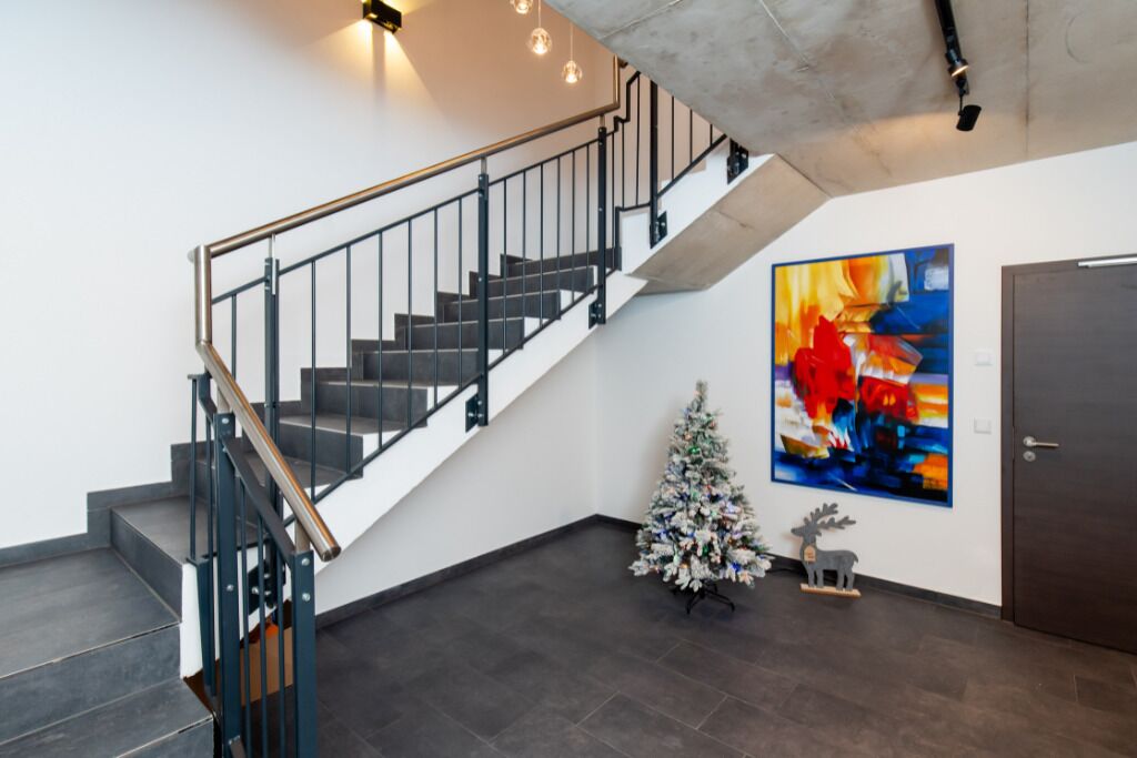 A staircase with a metal railing, a white wall, a colorful abstract painting, a small Christmas tree, and a door.