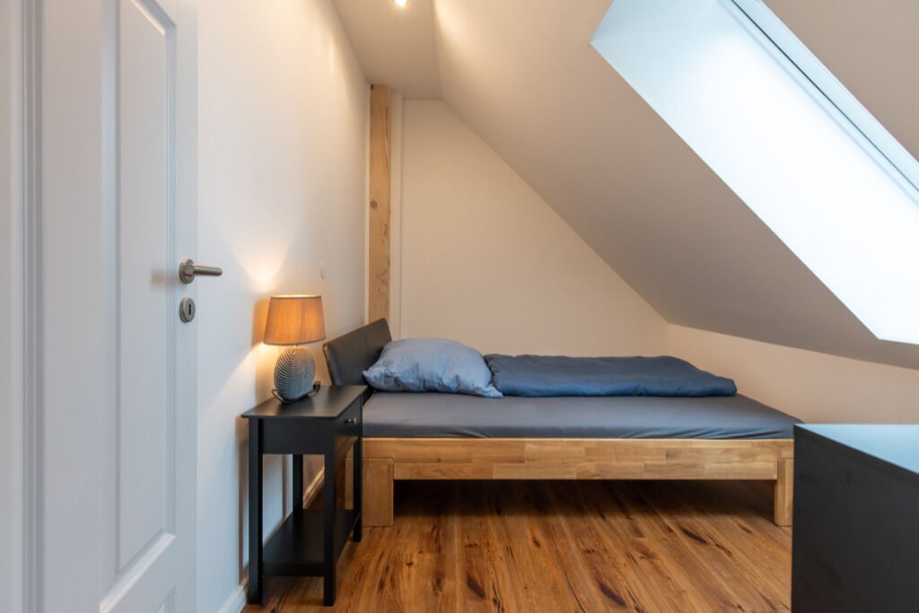 A small, cozy bedroom with a sloped ceiling, a bed with blue bedding, a bedside table with a lamp, and wooden flooring.
