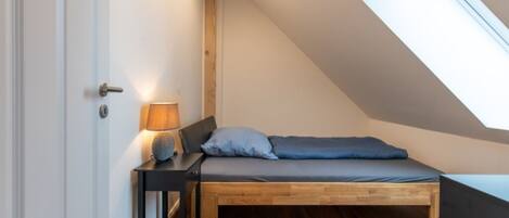 A small, cozy bedroom with a sloped ceiling, a bed with blue bedding, a bedside table with a lamp, and wooden flooring.
