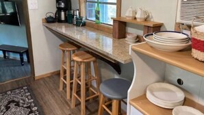 A kitchen with a wooden countertop, bar stools, a window with a view, and shelves with kitchenware. - Bluebird Nest (San Marcos)