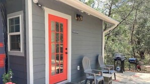 A small house with a red door, two Adirondack chairs, a barbecue grill, and a patio area. - Bluebird Nest (San Marcos)