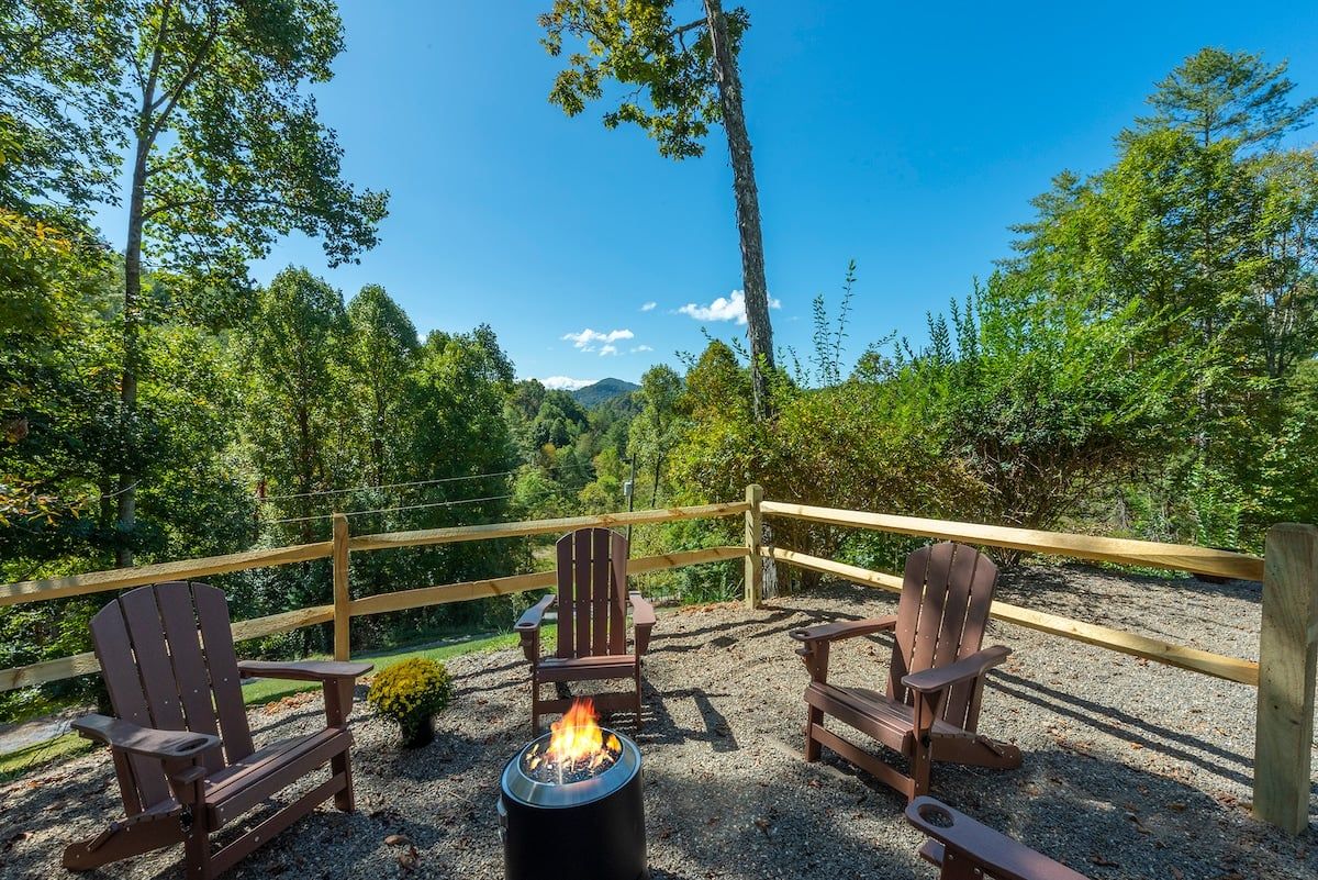 Eine Terrasse mit Adirondack-Stühlen und einem Feuerkorb, umgeben von einem Holzzaun und dichtem Wald.