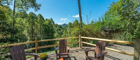 Eine Terrasse mit Adirondack-Stühlen und einem Feuerkorb, umgeben von einem Holzzaun und dichtem Wald.