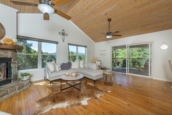 A spacious living room with a fireplace, a large sectional sofa, a wooden ceiling, and large windows offering a view of the outdoors.