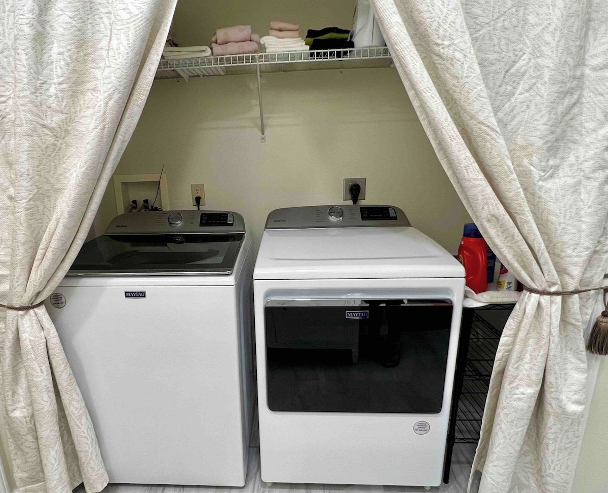 Un espace buanderie comprenant deux machines à laver Maytag, une étagère avec des serviettes pliées et une bouteille de lessive rouge.