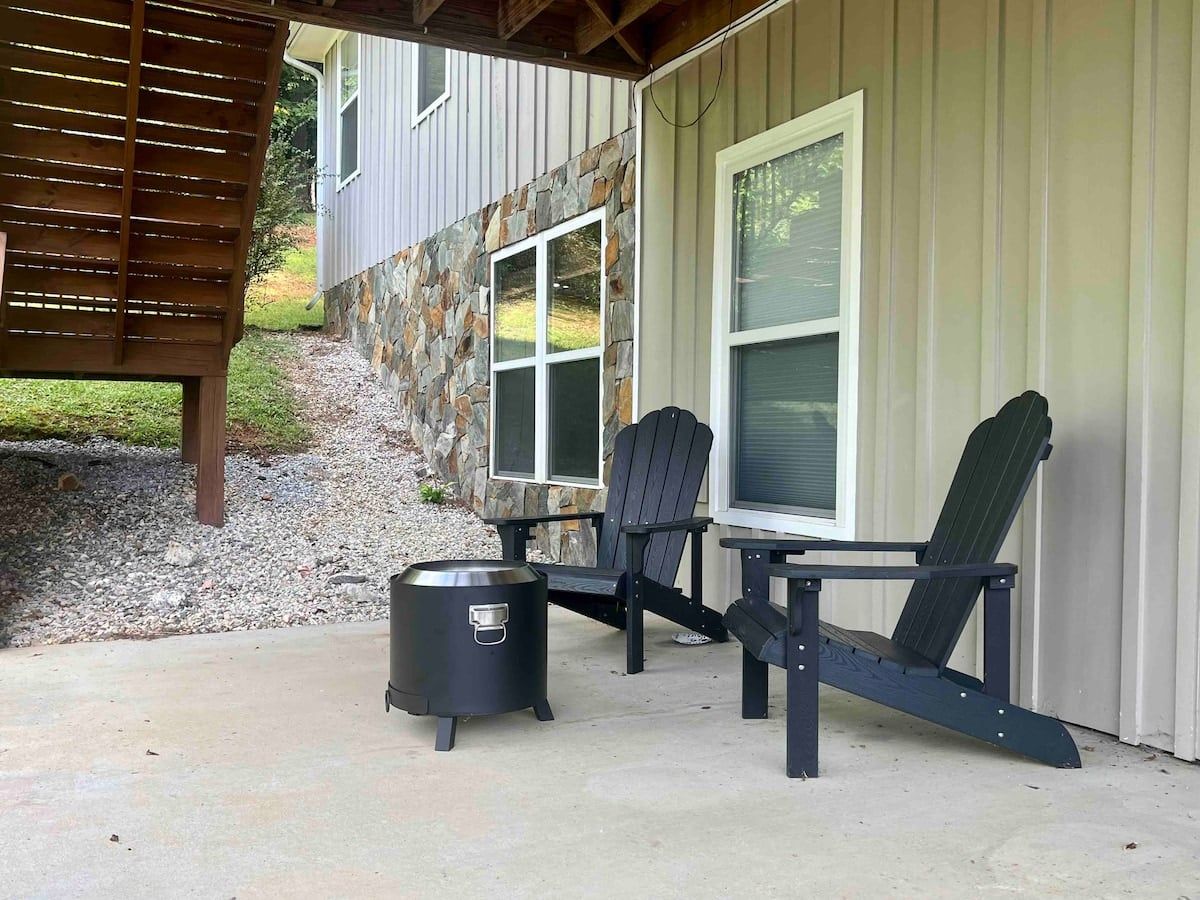 An outdoor seating area with Adirondack chairs and a grill, adjacent to a building with stone and siding exterior.