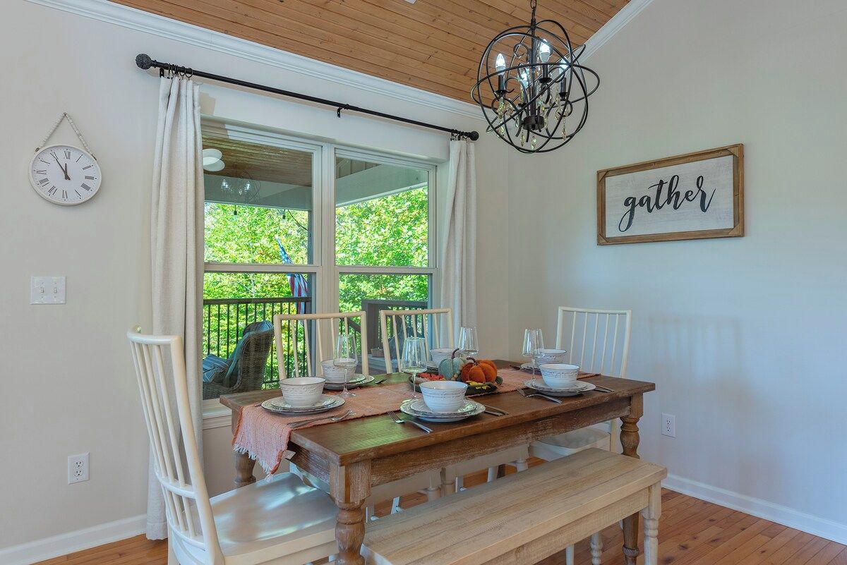 A dining area with a wooden table set for four, a chandelier, a 'gather' sign, and a view of greenery outside.