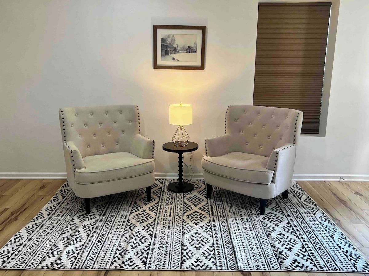A room with two armchairs, a small round table, a lamp, a patterned rug, and a framed picture on the wall.