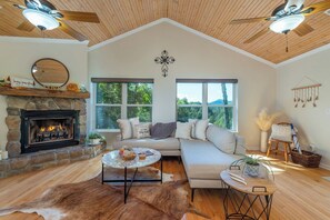 A cozy living room with a stone fireplace, a large window, a sofa, and a ceiling fan. - Mtn View, Fire Pit+Grill, WiFi! Nearby Town & AVL! (Burnsville)
