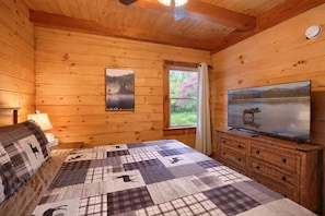 Ein Zimmer in einer Holzhütte mit einem Bett, einem Schminktisch, einem Fernseher und einem Fenster mit Blick ins Grüne.