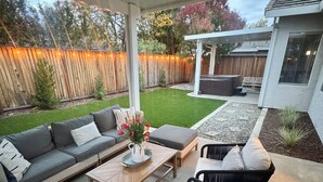 A covered patio area with a ceiling fan, outdoor seating including a sofa and chairs, a small table with a vase of flowers, and a wooden fence in the background. - Modern 4b 2bth Roseville Retreat with a spa Near Thunder Valley + The Grounds (Roseville)