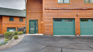A log cabin with two garage doors, a green door, and a small garden area with shrubs. - Lake Minocqua Condo w/ Shared Fire Pit! (Minocqua)
