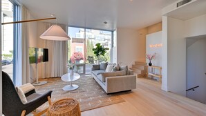 A modern living room with a sofa, armchair, and a TV on a stand. There is a large window with curtains, a floor lamp, and a staircase leading to an upper level. - Rooftop w/ Views: Modern Little Italy Townhome (San Diego)
