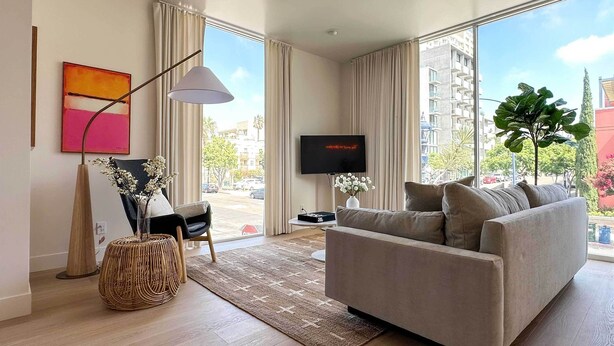 A modern living room with a sofa, a TV, a floor lamp, and a large window offering a city view. - Rooftop w/ Views: Modern Little Italy Townhome (San Diego)