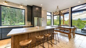 A modern kitchen with a large island, bar stools, and a view of the forest through large windows. - Modern Turnerville Cabin w/ Hot Tub & Scenic Views (Turnerville)