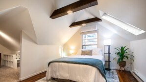 A bedroom with a sloped ceiling, a bed with a white and blue bedspread, a bedside table with a lamp, a potted plant, and a window with blinds.