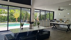 Une cuisine moderne dotée d’un grand îlot central, de tabourets de bar noirs et offrant une vue sur une piscine et un jardin tropical.