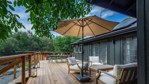 Una terraza de madera con mobiliario exterior, un paraguas grande y una vista a árboles y una casa al fondo.