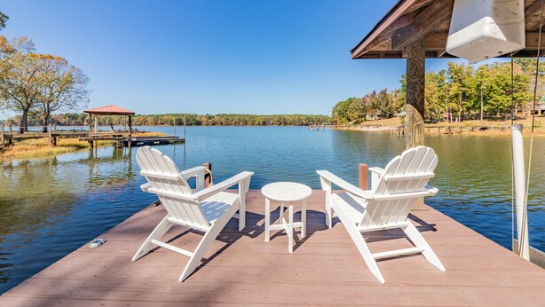 A wooden deck with two white Adirondack chairs and a small round table, overlooking a calm lake with a dock and a gazebo in the background.