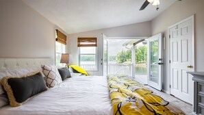 A bedroom with a large bed, a bench with a yellow and black pattern, a lamp, and a view of a balcony with a railing.