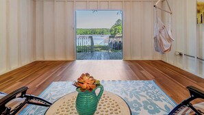 A room with a wooden floor, a view of a dock and water, a hanging hammock, and a table with a vase of flowers.