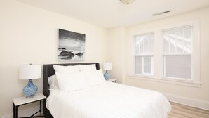 Une chambre à coucher avec un lit, deux tables de chevet, un tableau et une fenêtre avec des stores.