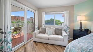 A bedroom with a balcony, a sofa, a lamp, and a view of buildings and trees.