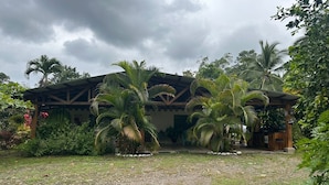 A building with a wooden structure and a covered entrance, surrounded by palm trees and greenery. - Riverside Lodge (Las Horquetas)