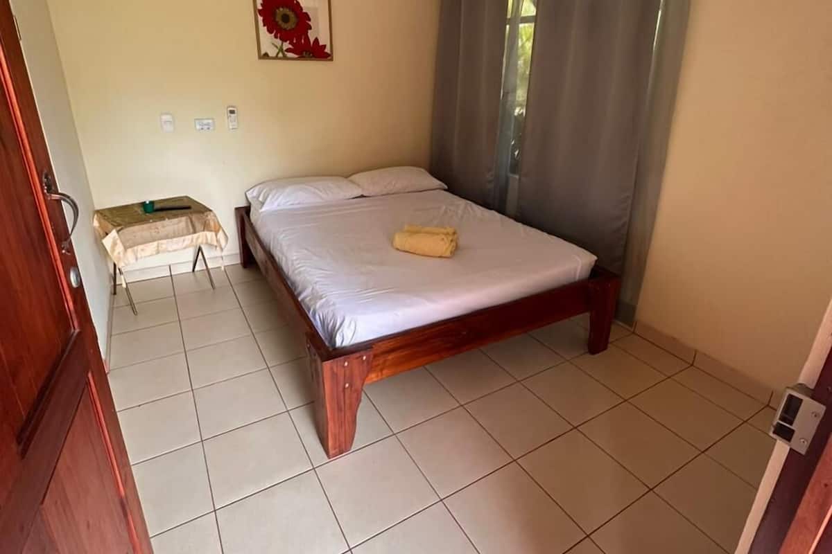 A bedroom with a wooden bed, a small bedside table, a wall-mounted air conditioner, and a window with curtains.