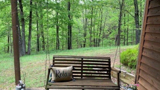 Un banco de madera con un cojín que dice “Relax” en un porche con vista a un bosque.