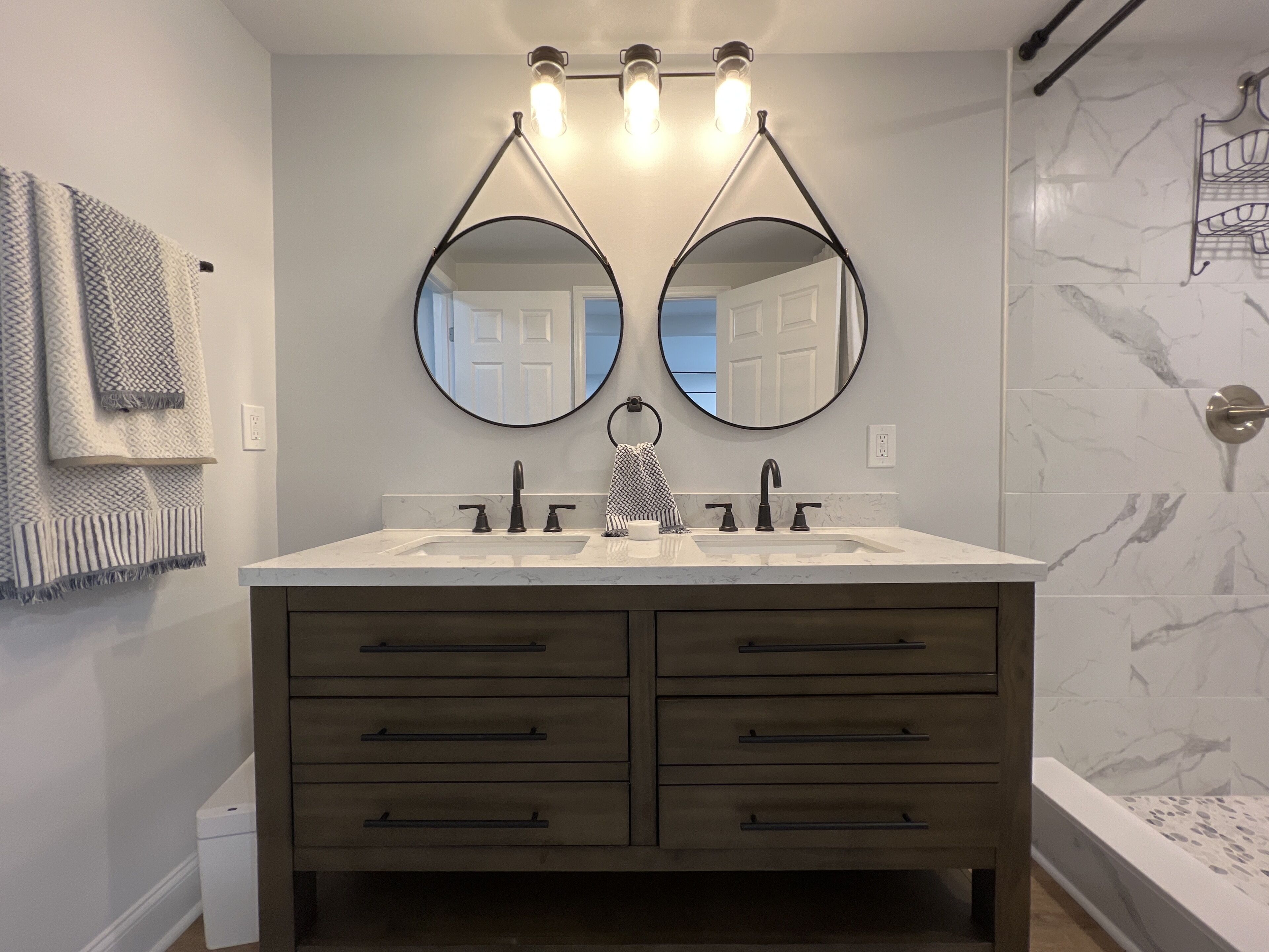 A bathroom with a double sink vanity, two round mirrors, and a shower area with a marble wall.