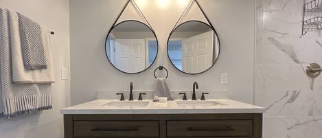 A bathroom with a double sink vanity, two round mirrors, and a shower area with a marble wall.