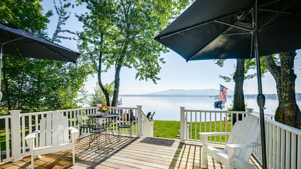 Una terraza de madera con barandillas blancas, mobiliario exterior y vista a un lago.