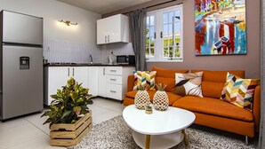 Cozinha moderna com armários brancos, uma geladeira e micro-ondas. Há um cesto de madeira com plantas, um sofá laranja com almofadas estampadas e uma mesa de centro branca. Um quadro colorido está na parede.