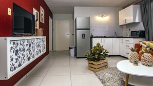 Cozinha moderna com armários brancos, uma geladeira de aço inoxidável e um micro-ondas. Há uma televisão em um suporte, uma lixeira e uma planta em vaso no chão.