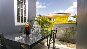 Mesa de vidro com cadeiras, um jarro de vidro e um copo com bebida, tudo diante de uma janela com vista para um edifício amarelo e uma planta.