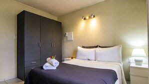 Cama bem arrumada com um cobre-leito escuro, mesa de cabeceira com abajur, e guarda-roupa com interruptor de luz na parede.