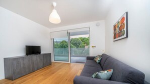 Una sala de estar moderna con un sofá gris, un televisor de pantalla plana sobre un mueble gris, un ventanal con vistas a la vegetación y una lámpara de techo.