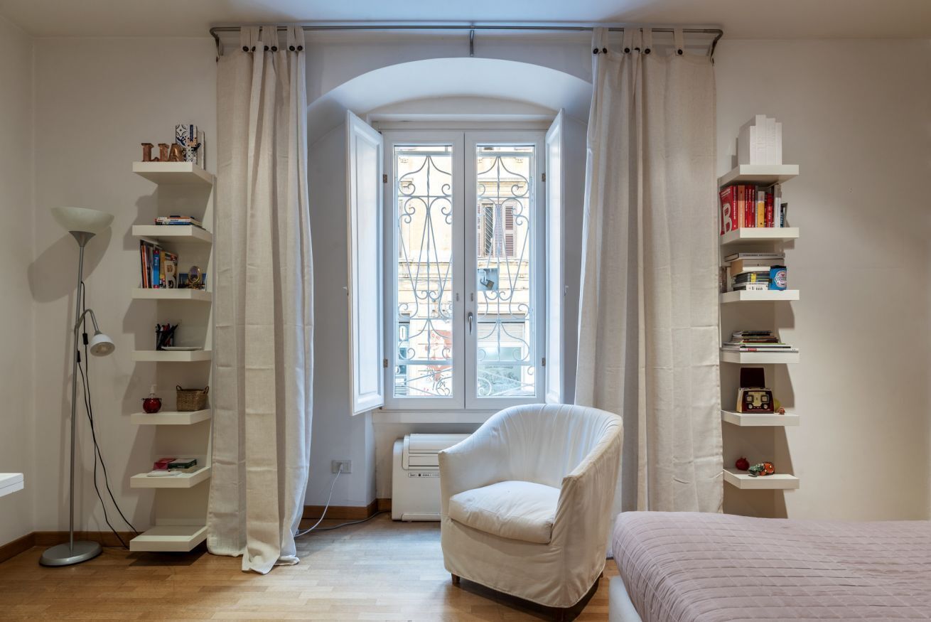 Ein Zimmer mit Fenster, einem Sessel, einem Bett und Regalen mit Büchern und Dekoration.