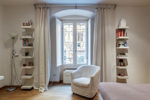 Ein Zimmer mit Fenster, einem Sessel, einem Bett und Regalen mit Büchern und Dekoration.