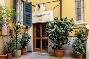 Entrada de un edificio con un letrero que dice 'SCALA C', plantas en macetas y una puerta con paneles de vidrio.
