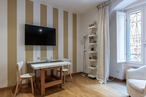 Ein Zimmer mit an der Wand montiertem Fernseher, einem Holztisch mit Stühlen, einem Bücherregal und einem Fenster mit Vorhang.